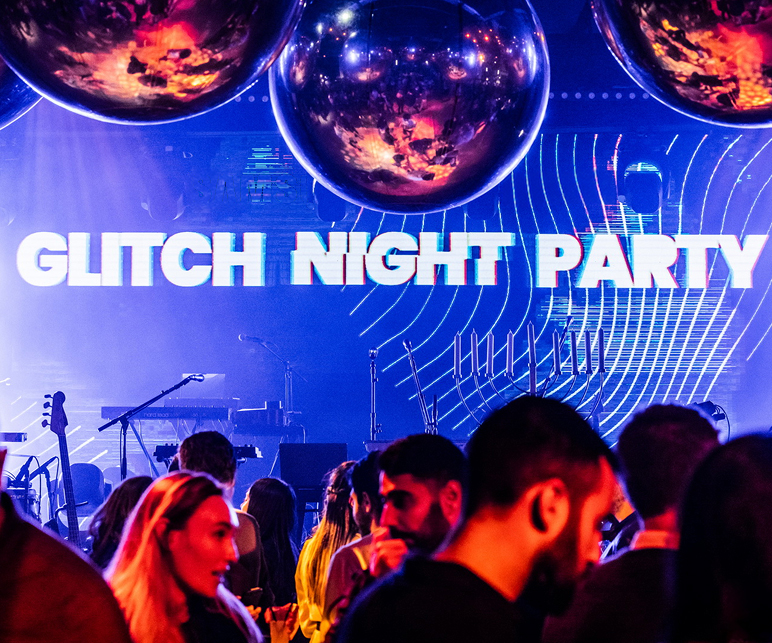 “Glitch Night Party” event with a large blue LED stage backdrop, hanging mirrored disco balls, live music equipment, and guests gathered in the foreground under vibrant blue, pink, and red lighting.