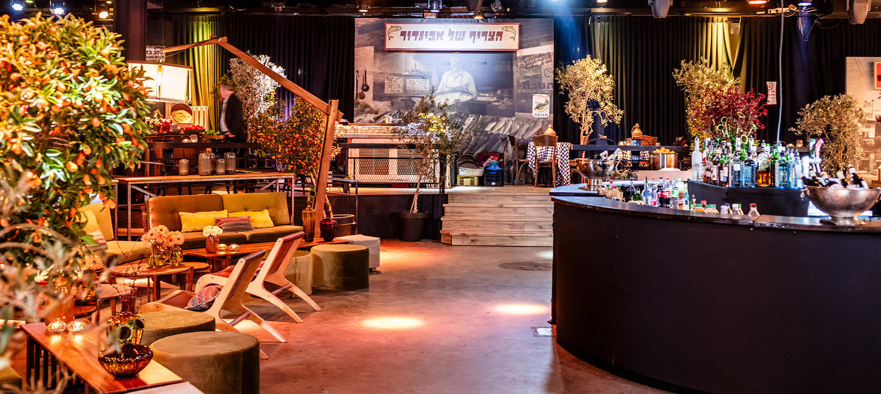 event lounge and bar setup with olive and citrus trees, warm lighting, rustic decor, and a market-style backdrop.